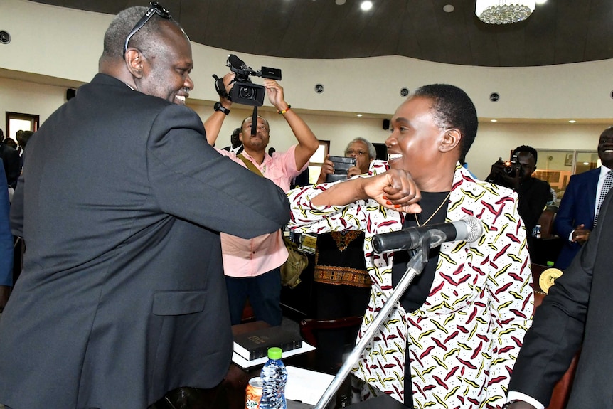 Riek Machar smiles as he elbow bumps with his wife as photographers lean in to take pictures