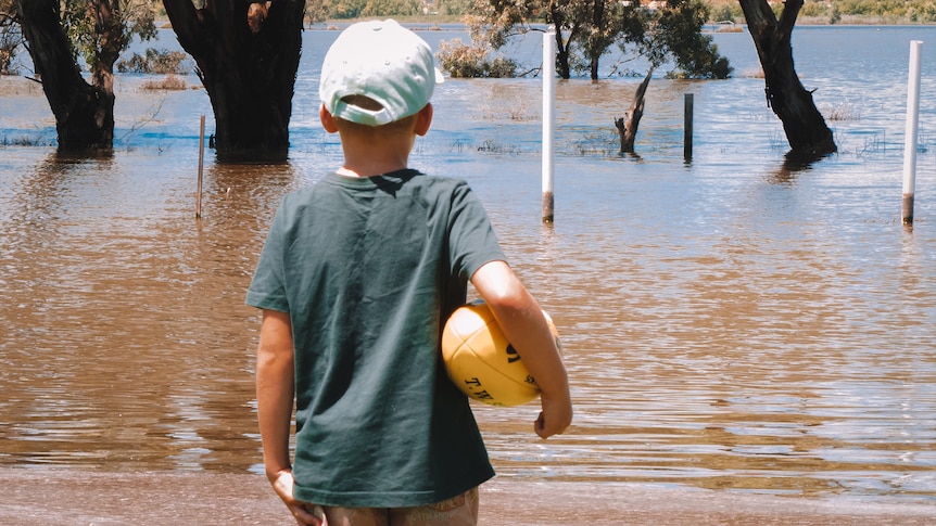 A little boy holding a football looks out over a flooded field.