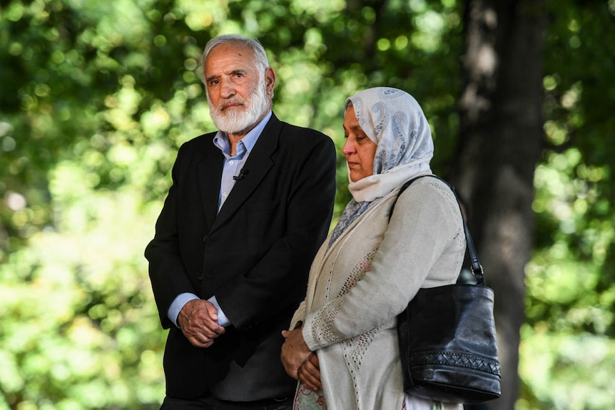 A man and woman stand in front of a row of green trees.