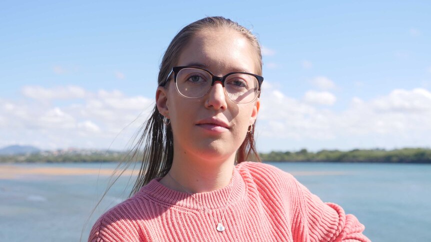 A woman wearing glasses and a pink jumper standing in front of a river
