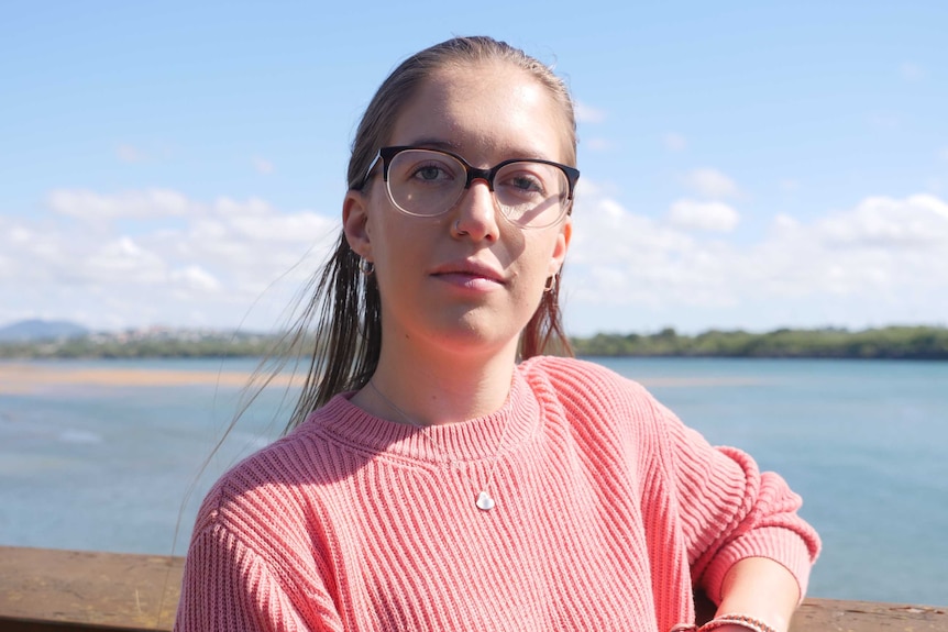 A woman wearing glasses and a pink jumper standing in front of a river