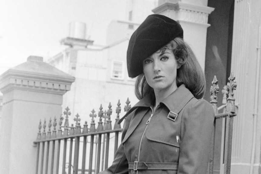 A woman in a beret and trench coat standing on the street and holding her thumb out as a hitchhiker would.