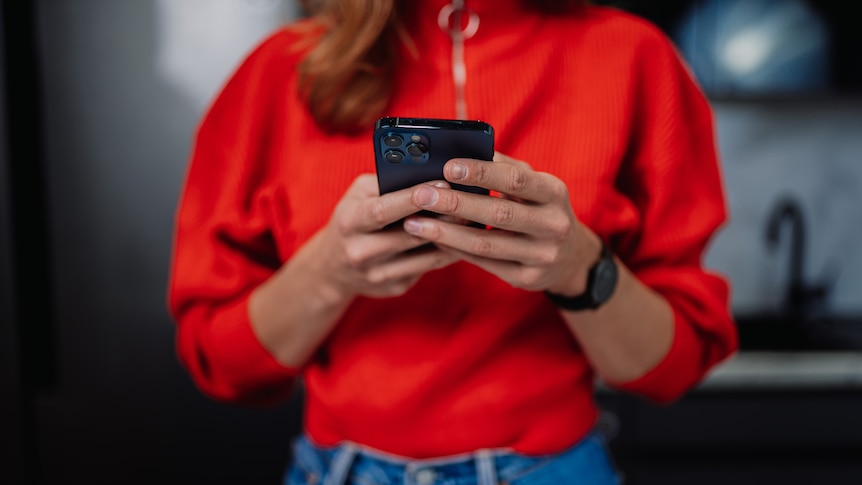 A woman in a bright red sweater and blue jeans texts on her smartphone