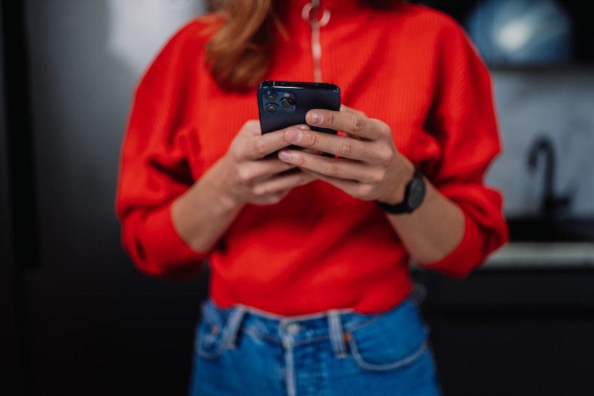 A woman in a bright red sweater and blue jeans texts on her smartphone