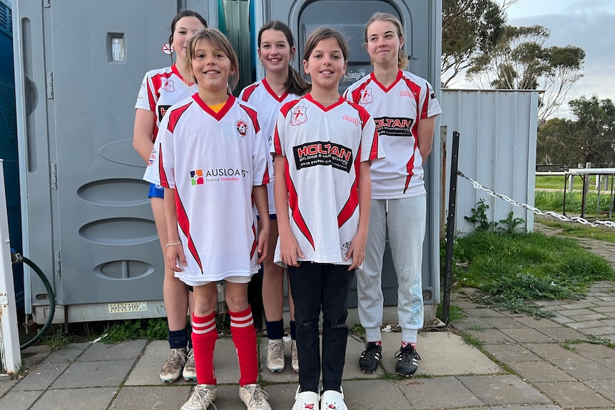 Five young female soccer players stand in front of a group of portaloos