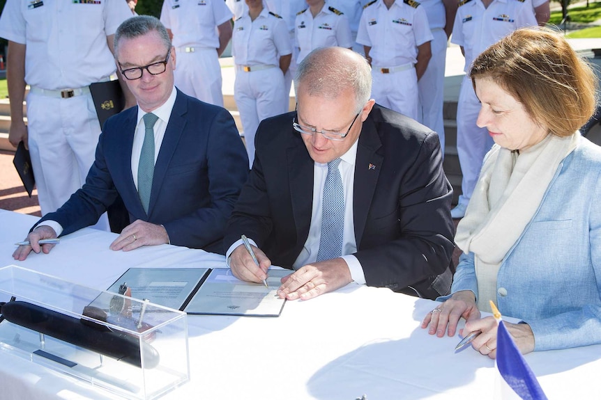 Scott Morrison signs a document with Christopher Pyne and Florence Parly sitting alongside him. Naval officers stand behind them