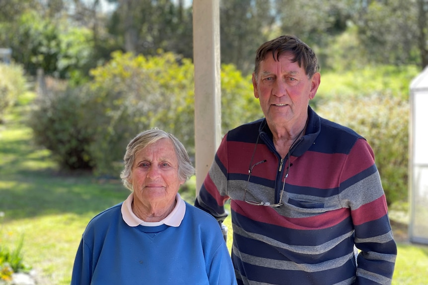 A woman in a blue jumper and a man in a striped jumper look at the camera under a carport