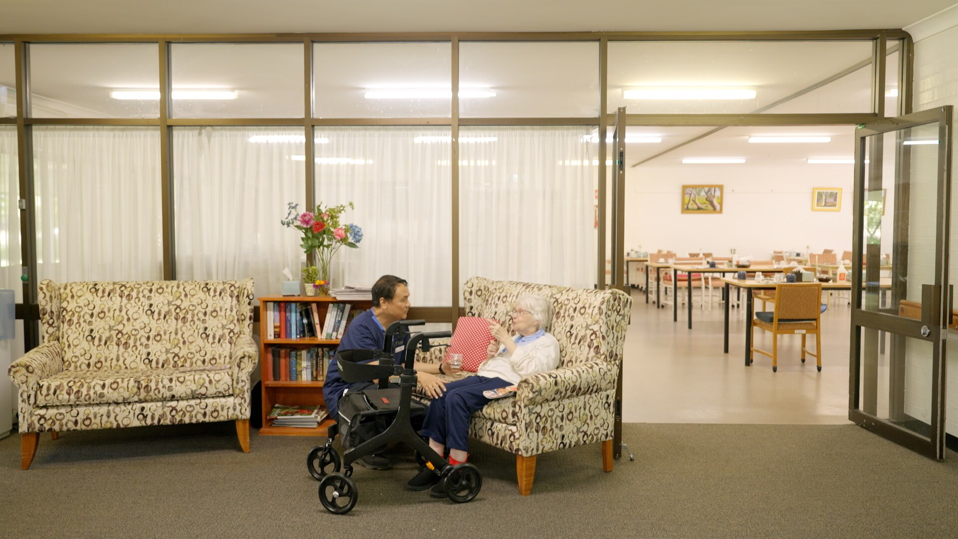 Male healthcare worker squats next to elderly woman seated on couch in the foyer of an aged care facility