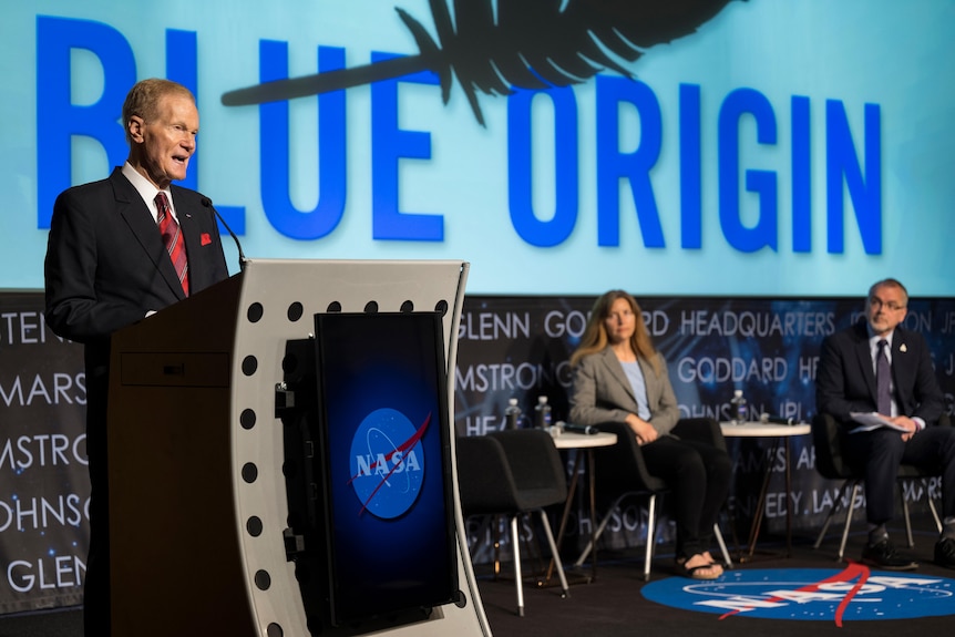 Suited man stands at a lecturn, featuring a NASA logo, on a stage where two other people are seated