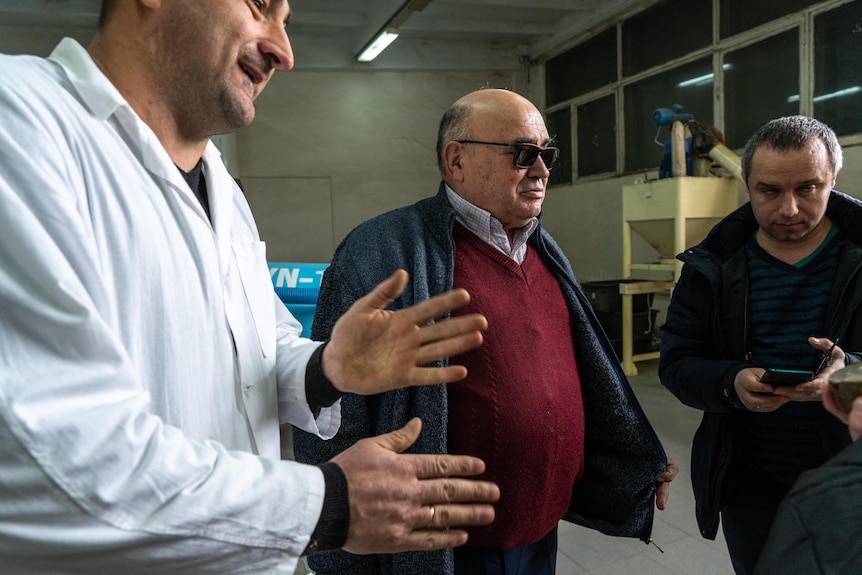 A man in a labcoat speaks to two other men inside a biofuel lab.