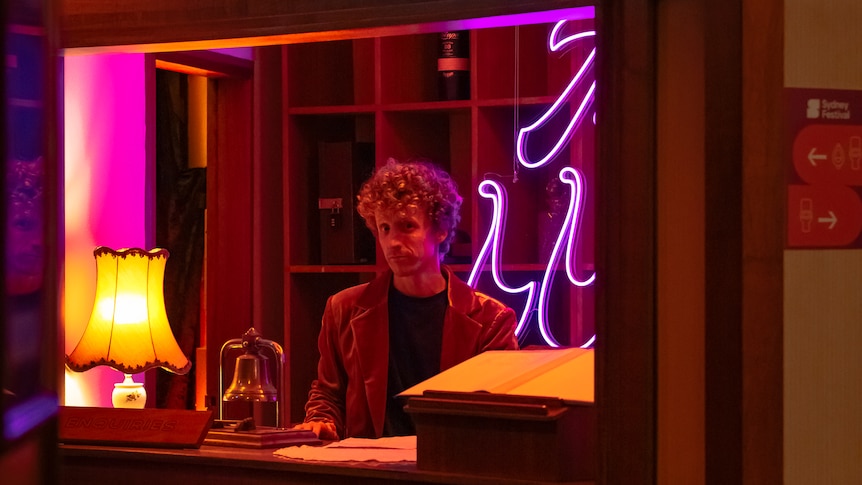 A blonde curly haired man stands behind the front desk of a hotel warmly lit by a 70s style orange lamp and a neon pink sign.