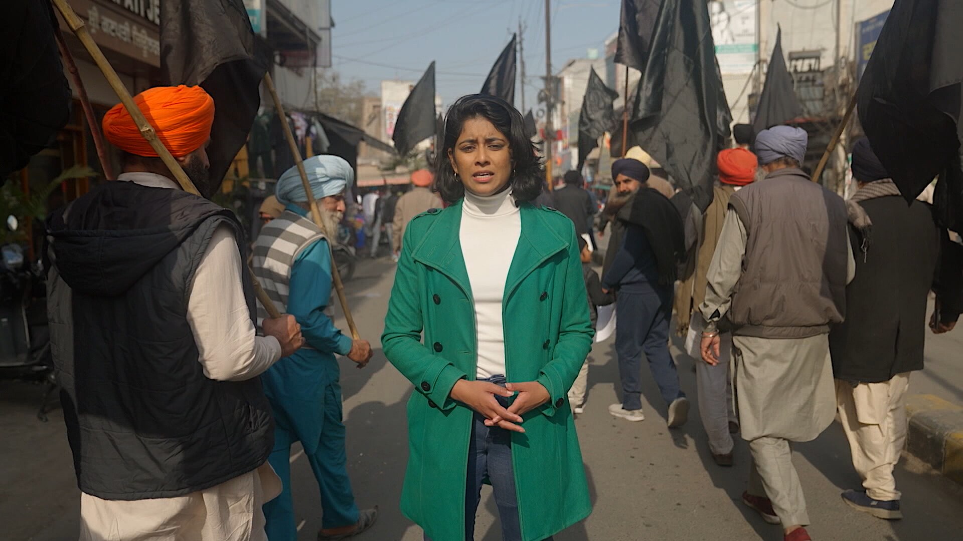 A woman in a green coat talking to the camera as a crowd of men wearing turbans walk by her