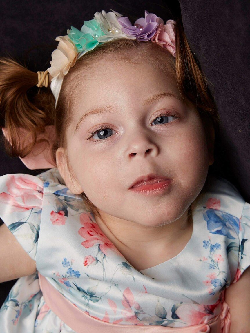 Portrait of Tahlea, two-and-a-half-years old, she has pigtails and wears a rainbow flower headband and a floral dress.