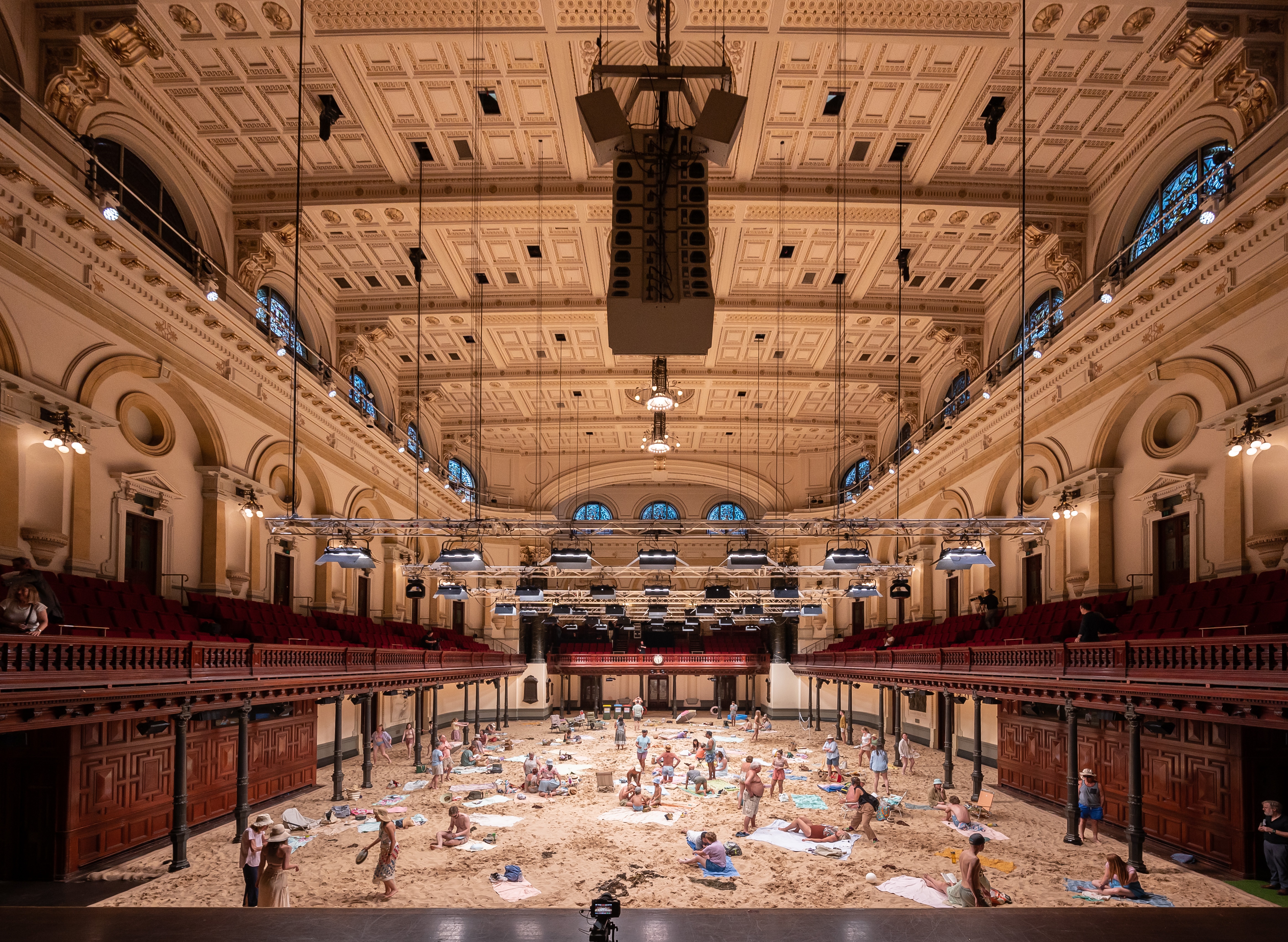 Sand a people dressed as beach-goers cover the flooring of a large ornate and neoclassical-designed hall.