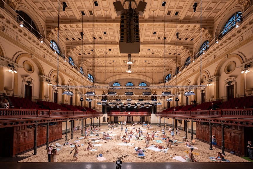 Sand a people dressed as beach-goers cover the flooring of a large ornate and neoclassical-designed hall.