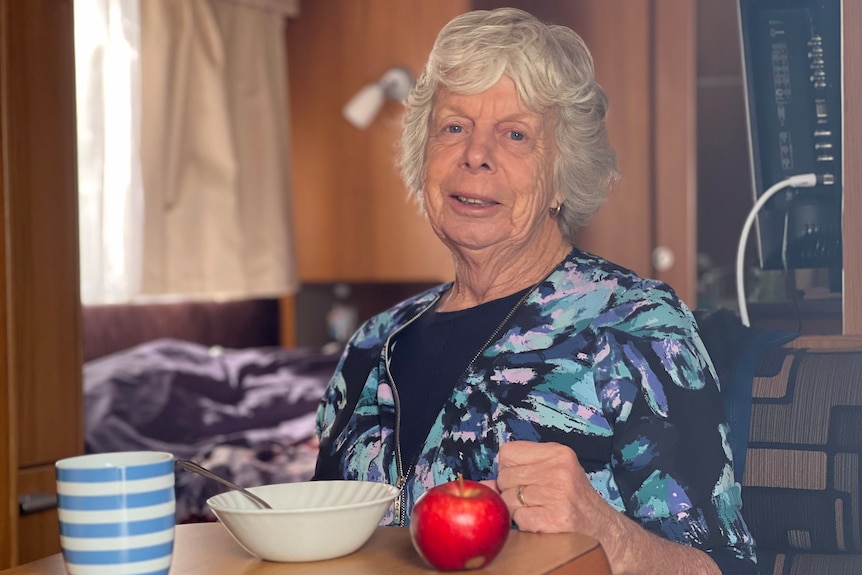 A woman wearing a blue and black top sits at a table with a mug, bowl and apple.