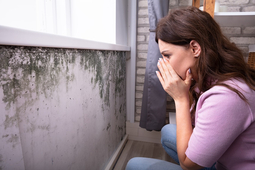 Woman covering her mouth and nose with her hands as she looks at mould on a wall in a home.