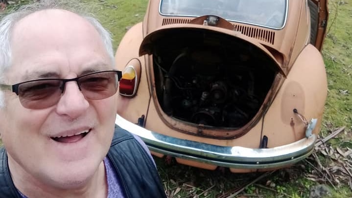 Old man taking selfie with an old honey brown VW beetle behind him.