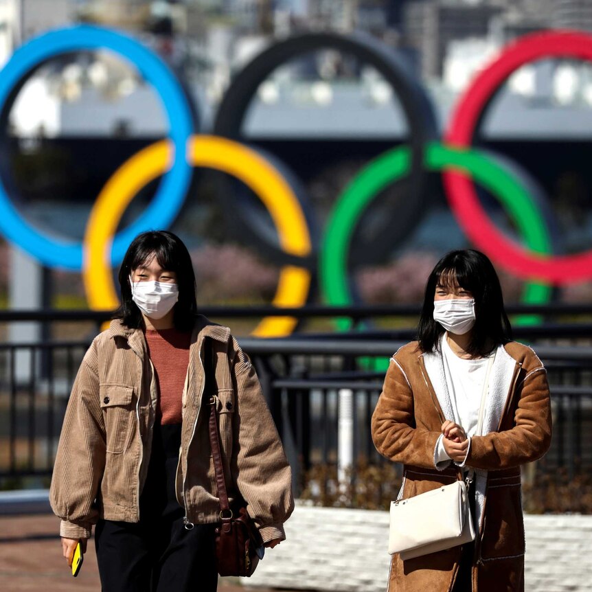 Two women in surgical masks walking in front of a sculpture of the Olympic rings