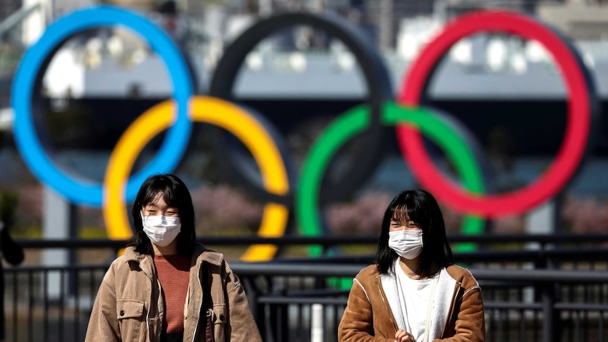 Two women in surgical masks walking in front of a sculpture of the Olympic rings