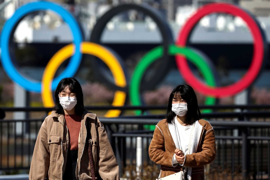 Two women in surgical masks walking in front of a sculpture of the Olympic rings