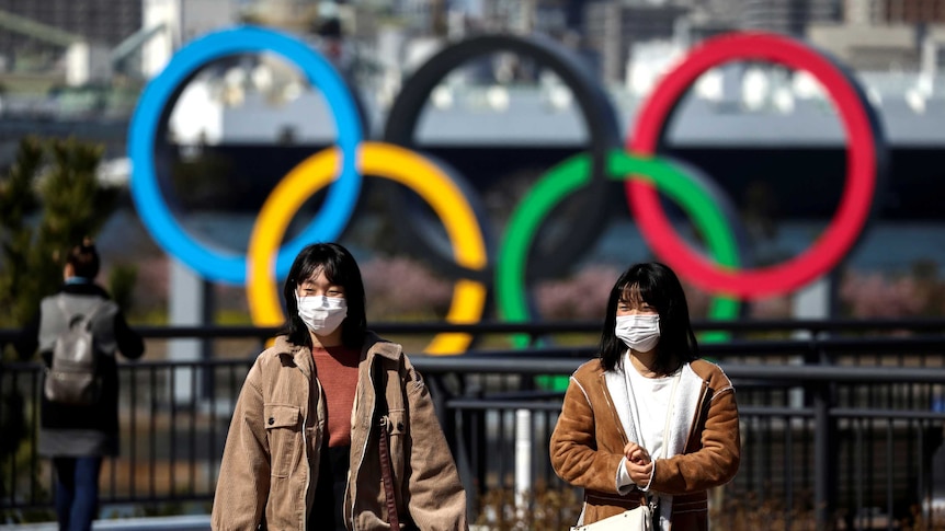 Two women in surgical masks walking in front of a sculpture of the Olympic rings