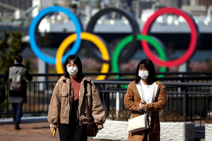 Two women in surgical masks walking in front of a sculpture of the Olympic rings