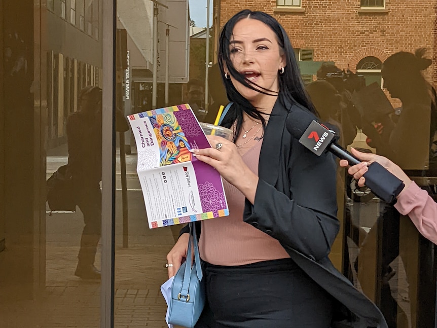 A woman with a drink and book speaks to media outside a courthouse