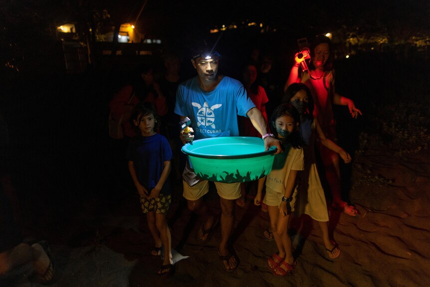 A man carries turles in a bucket in the dark while wearing a torch and surrounded by other people. 