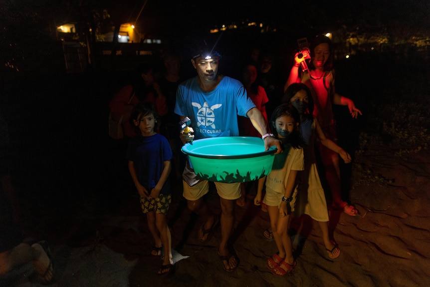 A man carries turles in a bucket in the dark while wearing a torch and surrounded by other people. 