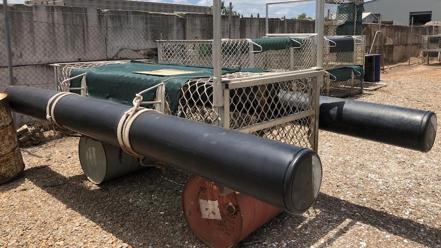 Large aluminium cage with floating pontoons either side sitting in a gravel yard.