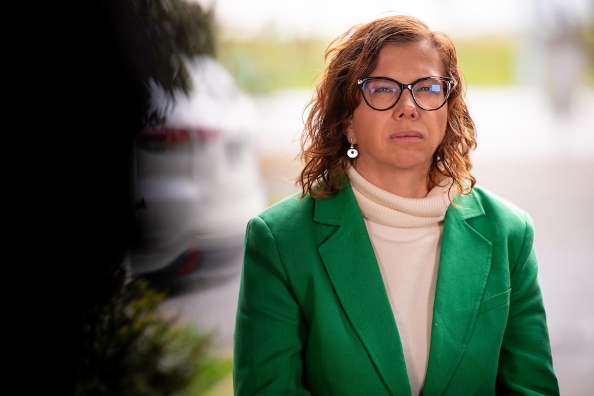 A woman wearing a green jacket and a white turtleneck with brown hair and glasses looks serious