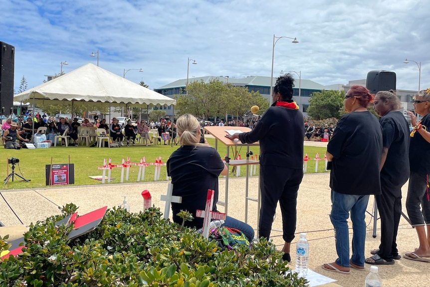 A row of women stand in front of a seated crowd addressing them at Edith Cowan Square in Geraldton. under a blue sky.