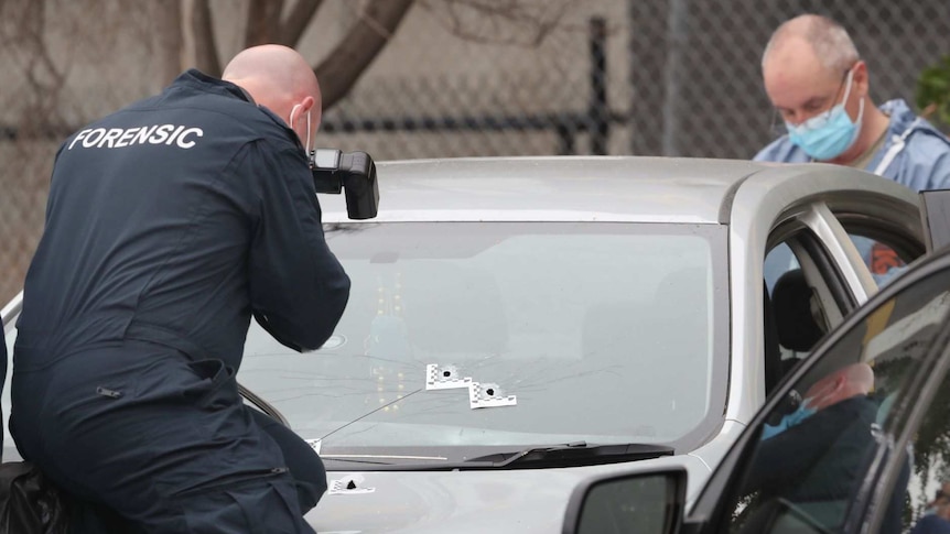 A police forensics officer sits on the bonnet of a silver car, taking photos of three bullet holes in the windscreen and bonnet