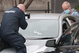 A police forensics officer sits on the bonnet of a silver car, taking photos of three bullet holes in the windscreen and bonnet