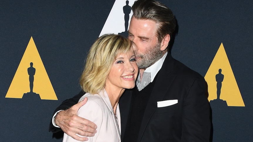 A fair-haired woman with a wide smile and sparkling eyes is embraced by a bearded man with dark hair who is kissing her head