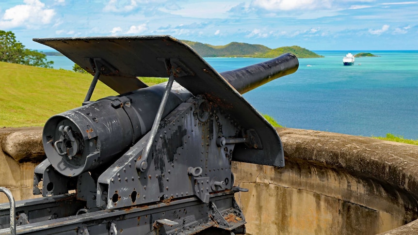 A large artillery gun is pointed out to sea where a cruise ship lies at anchor.