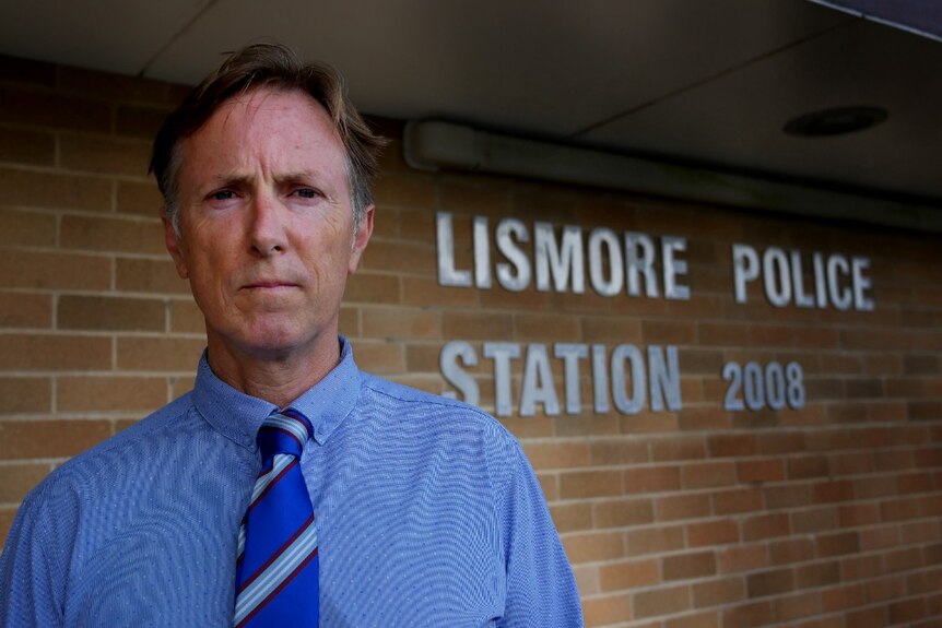 A man in a blue shirt with a tie in front of a sign that says 'Lismore Police Station 2008'.