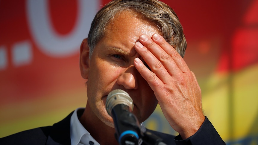 A man in a suit holds his hand over his eyes from the sun as he speaks into a microphone