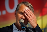 A man in a suit holds his hand over his eyes from the sun as he speaks into a microphone