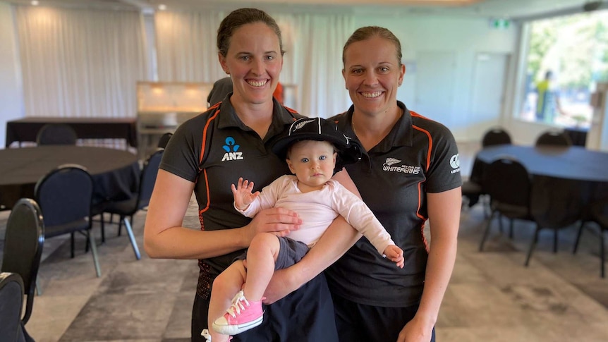 Two female cricketers smile at the camera, with one holding a baby in her arms.