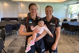 Two female cricketers smile at the camera, with one holding a baby in her arms.