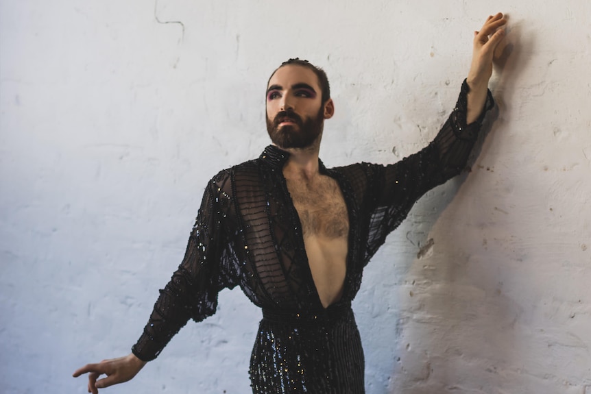 Matthew Pope poses in full makeup in a sequined suit and his arm against a white wall. 