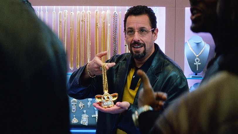 A man in leather jacket with black beard, short curly hair presents bedazzled Furby necklace to two figures in jewellery store.