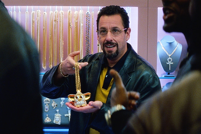 A man in leather jacket with black beard, short curly hair presents bedazzled Furby necklace to two figures in jewellery store.