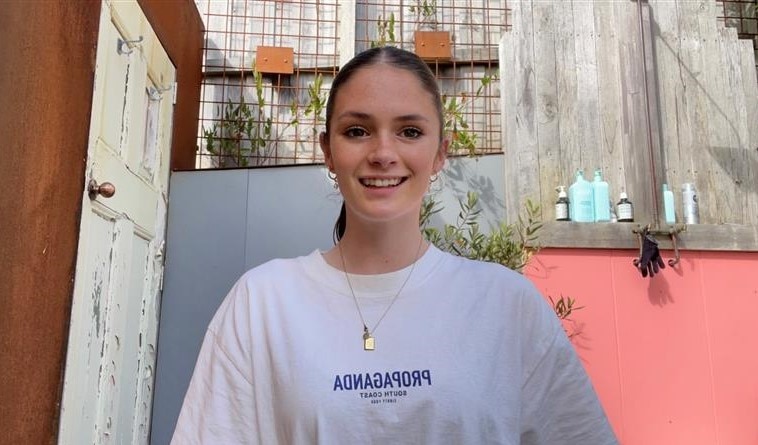 young girl with long brown hair tied up in a ponytail smiles at camera wearing a white t shirt
