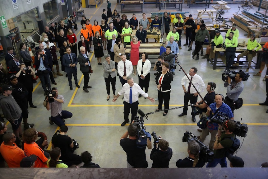 Bill Shorten talking in an industrial room with his arms outstretched.He's surrounded by his colleagues, media and TAFE students