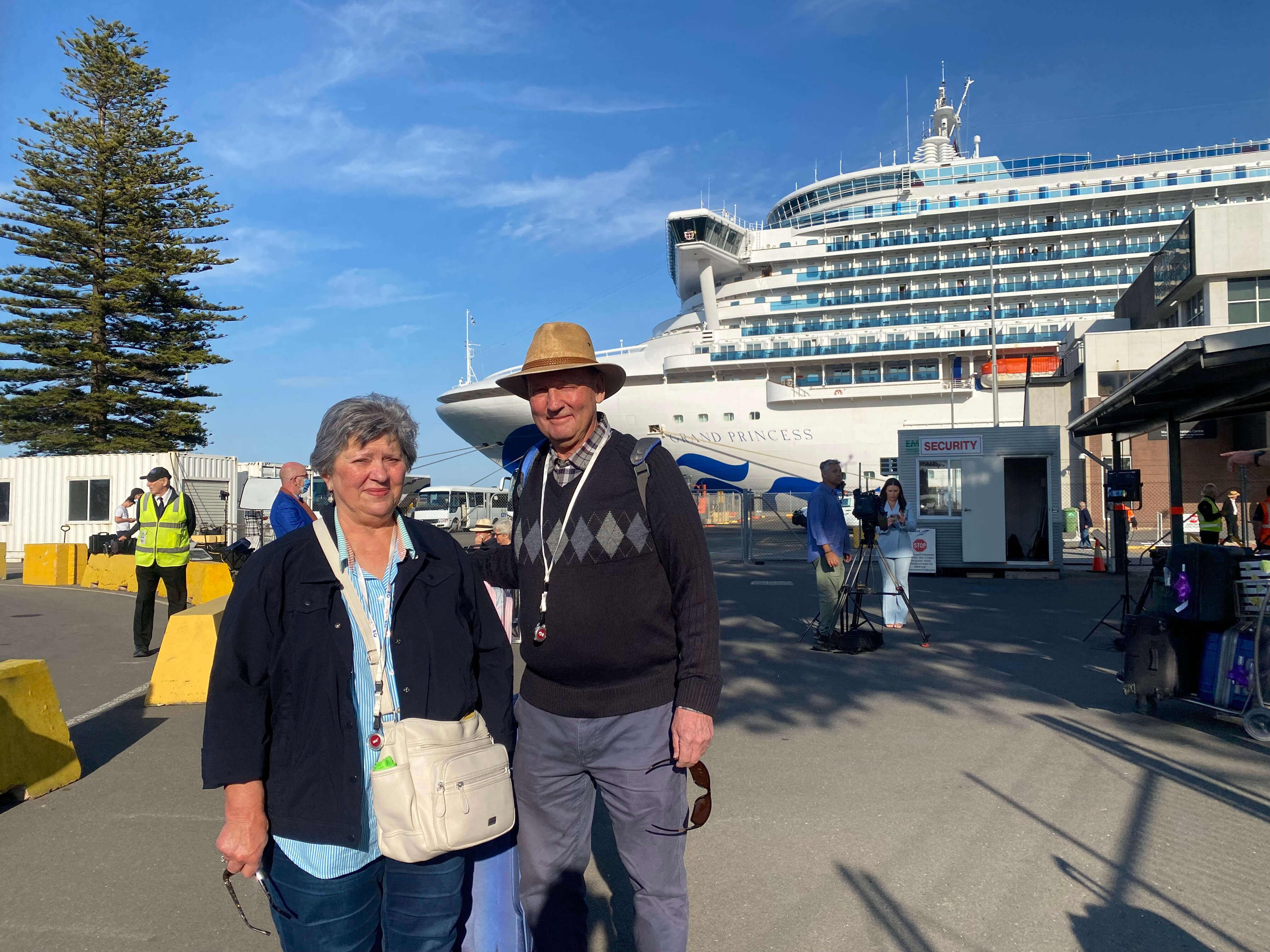A woman and a man, with their bags, at a harbour with a large cruise ship behind