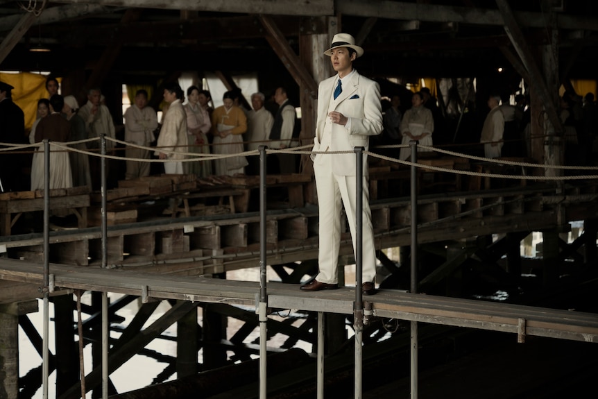 A rich Korean man stands in a white suit stands looking out at the view, behind him is a crowded, dark and dank marketplace.