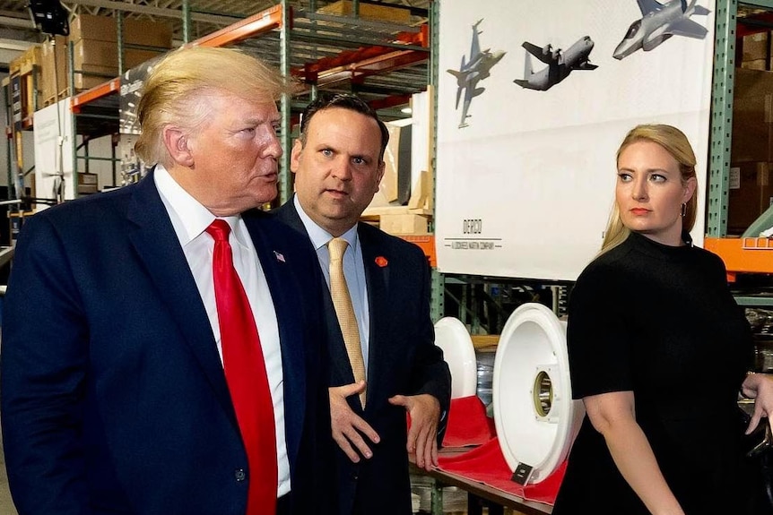 Emma Doyle walks alongside Donald Trump and another man in a warehouse. A poster behind her shows aeroplanes.